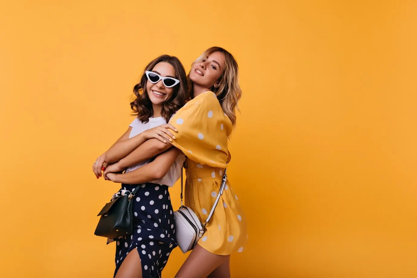 Ein fröhliches Mädchen mit weißer Vintage-Sonnenbrille genießt Freizeit mit einer befreundeten Person. Glamouröse, gut gekleidete Frauen umarmen sich vor gelbem Hintergrund.